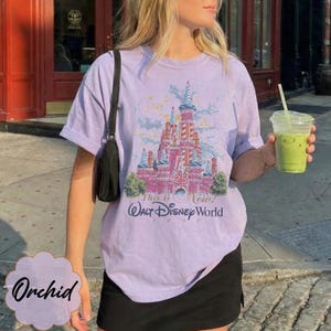 K&ouml;nnte beinhalten: Ein hellviolettes Kurzarm-T-Shirt mit einer Grafik eines rosafarbenen Schlosses und dem Text "25 This is Now! Walt Disney World 1997". Das T-Shirt wird mit einer schwarzen Umh&auml;ngetasche und einem schwarzen Rock kombiniert, w&auml;hrend ein Becher gehalten wird.
