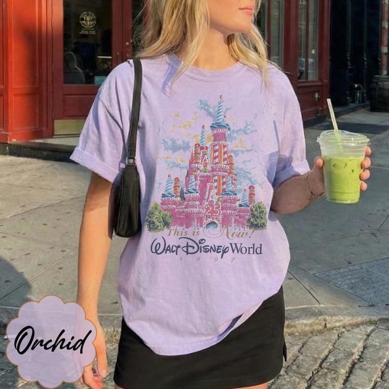 K&ouml;nnte beinhalten: Ein hellviolettes Kurzarm-T-Shirt mit einer Grafik eines rosafarbenen Schlosses und dem Text "25 This is Now! Walt Disney World 1997". Das T-Shirt wird mit einer schwarzen Umh&auml;ngetasche und einem schwarzen Rock kombiniert, w&auml;hrend ein Becher gehalten wird.