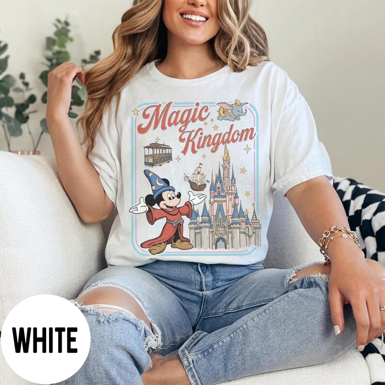 May include: White t-shirt with a retro graphic of Mickey Mouse as a wizard, a castle, and the words "Magic Kingdom." The design includes a tram and a ship. The shirt is worn by a person wearing blue jeans.