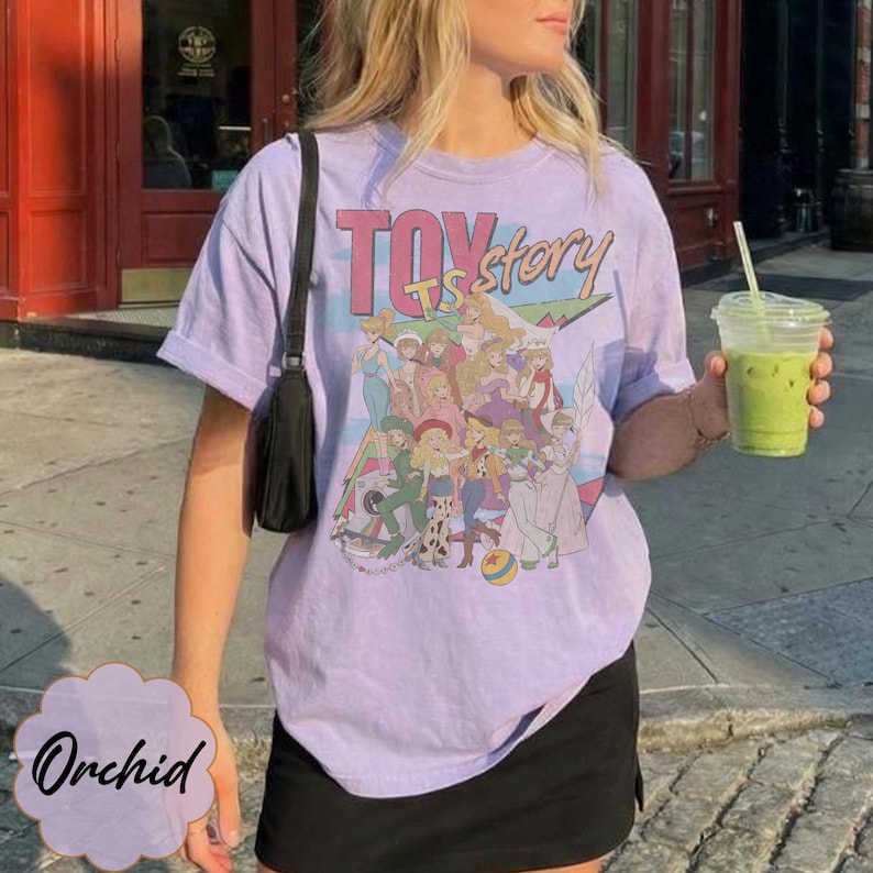 May include: A light purple short-sleeve t-shirt featuring a graphic print of various animated characters and the text Toy Story. The shirt is paired with a black skirt and a black shoulder bag, while a clear plastic cup with a green drink is held.