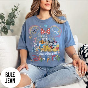 May include: A blue t-shirt featuring a colorful graphic of Disney characters, including Mickey and Minnie Mouse, with a castle and fireworks. The text "my FIRST Disney TRIP" is displayed below the graphic. The shirt has short sleeves.