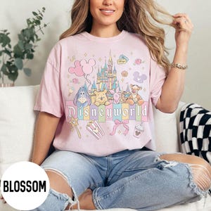 May include: A light pink t-shirt featuring a colorful graphic of Disney characters and a castle. The word "Disneyworld" is printed across the front. The shirt is worn by a person wearing blue jeans.