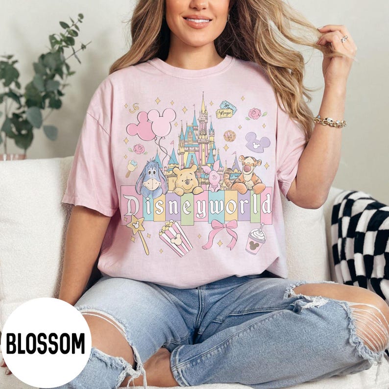 May include: A light pink t-shirt featuring a colorful graphic of Disney characters and a castle. The word "Disneyworld" is printed across the front. The shirt is worn by a person wearing blue jeans.