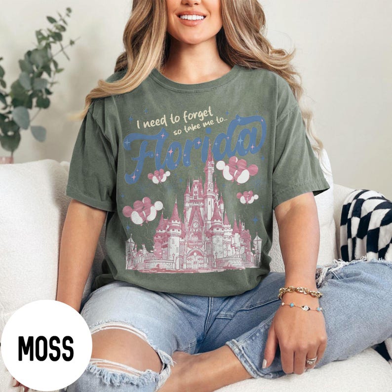 May include: Moss green t-shirt with the text "I need to forget so take me to Florida" in blue and pink. The shirt features a pink castle graphic with clouds. The model is wearing blue jeans.