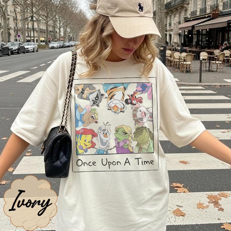 May include: Ivory-colored t-shirt featuring a colorful cartoon graphic with the text "Once Upon A Time." The graphic includes various animated characters. The shirt is paired with a beige baseball cap and a black purse with a chain strap.