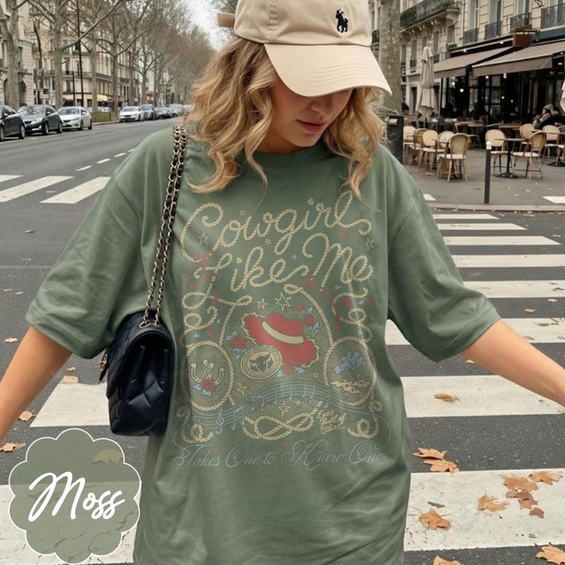 May include: A moss green short-sleeve t-shirt featuring a graphic print with the text Cowgirl like Me and Takes One to Know One. The design includes a red cowboy hat, a rope border, musical notes, and stars. A beige baseball cap is worn above.