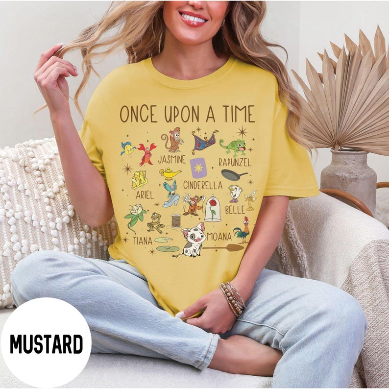 May include: Mustard-coloured t-shirt with the words "ONCE UPON A TIME" and various cartoon character illustrations. The shirt features characters like Jasmine, Cinderella, and Moana. The word "MUSTARD" is printed on the bottom left.