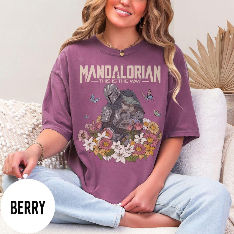 Puede incluir: Camiseta color bayas con un gr&aacute;fico del Mandaloriano y Grogu rodeados de flores y mariposas. El texto "MANDALORIAN - THIS IS THE WAY" est&aacute; impreso en la parte superior. La camiseta tiene mangas cortas y un corte holgado.