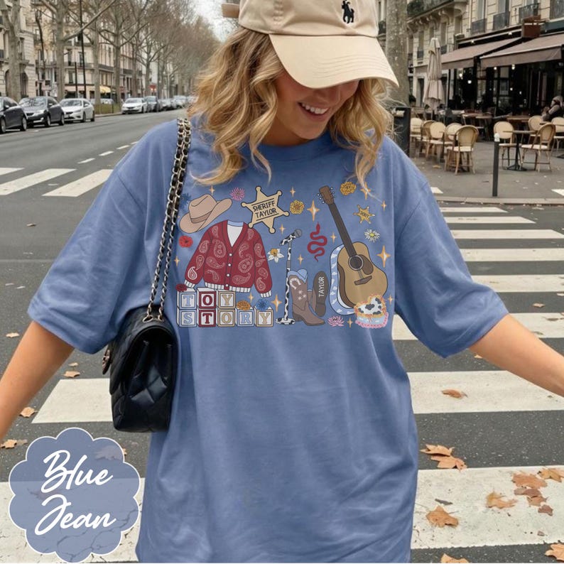 May include: A blue t-shirt featuring a graphic print of a cowboy hat, a red paisley cardigan, a sheriff badge labeled Sheriff Taylor, a microphone, a cowboy boot, an acoustic guitar, and blocks spelling Toy Story with a black shoulder bag.