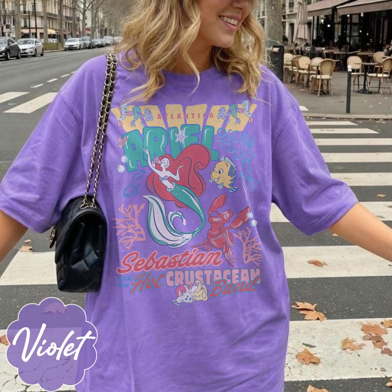 May include: Violet purple t-shirt featuring a graphic of Ariel, Sebastian, and Flounder from The Little Mermaid. The shirt has the words "Atlantica" and "Crustacean" printed on it. A black purse with a chain strap is visible.