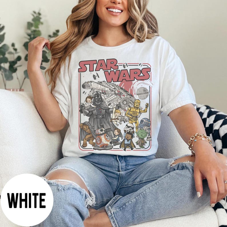 May include: White t-shirt featuring a Star Wars graphic. The design includes the Star Wars logo, Darth Vader, the Millennium Falcon, and other characters. The shirt is a classic crew neck style.