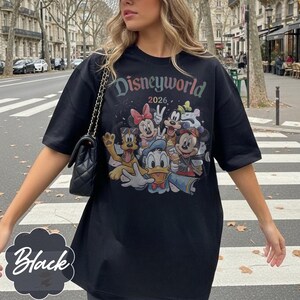 Pu&ograve; includere: T-shirt nero con una grafica colorata di personaggi Disney, tra cui Topolino, Minnie e Paperino. Il testo "Disneyworld 2026" &egrave; stampato sopra i personaggi. La maglietta &egrave; in stile casual.