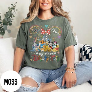 May include: Moss green t-shirt featuring a colorful Disney character design. The design includes a castle, Mickey and Minnie Mouse, and the text "my FIRST Disney TRIP". A red bow with white polka dots and the word "Name" are also featured.