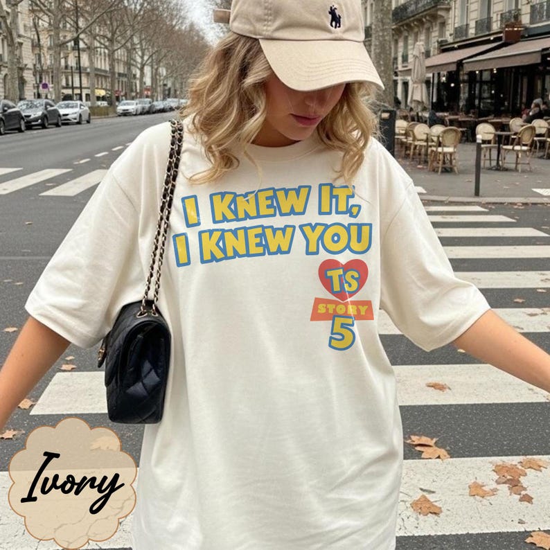 May include: An ivory short-sleeve t-shirt featuring the blue text I KNEW IT, I KNEW YOU, a red heart with the letters TS, and the word STORY above the number 5 in yellow. A black quilted shoulder bag with a gold chain strap is worn over the shoulder.