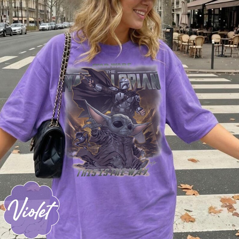 Puede incluir: Una camiseta violeta con un estampado gr&aacute;fico de The Mandalorian y Grogu. El texto dice Star Wars The Mandalorian y This Is The Way. La camiseta tiene mangas cortas y cuello redondo. Se ve un bolso de hombro negro acolchado con cadena.
