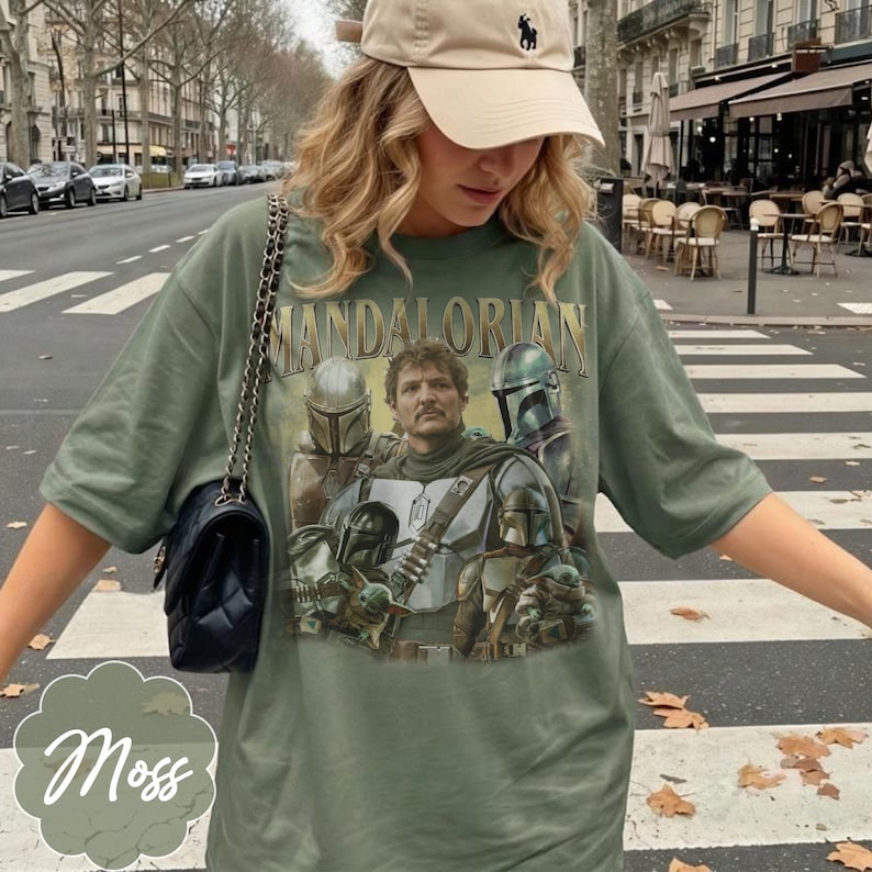Pu&ograve; includere: Una t-shirt verde oliva con una stampa grafica dei personaggi di The Mandalorian, tra cui una figura centrale con i baffi, varie figure corazzate con elmo e una piccola creatura verde. &Egrave; visibile anche una borsa a tracolla nera trapuntata.