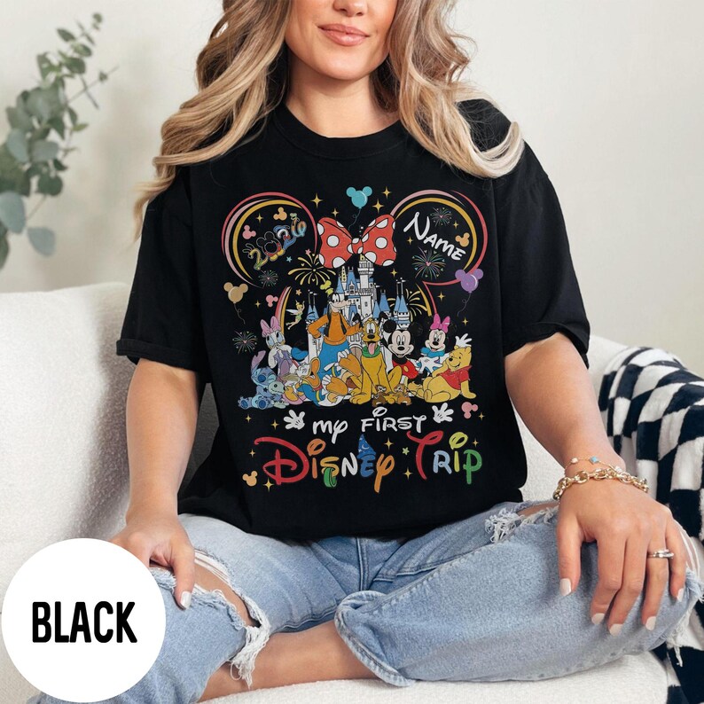 May include: Black t-shirt featuring a colorful graphic of Disney characters, a castle, and the text "my FIRST Disney TRIP." The design includes a large bow, fireworks, and the word "NAME" in a circle. The shirt is worn by a person.