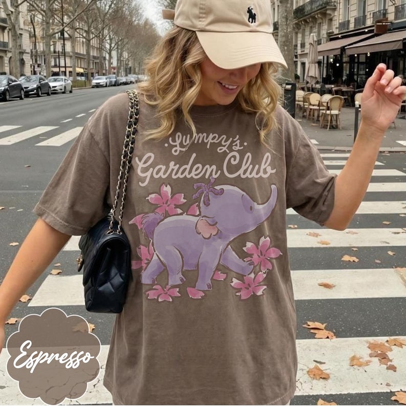 Op de afbeelding: Espresso-kleurig T-shirt met een paars olifantenontwerp en roze bloemen, met de tekst "Lumpy's Garden Club". Het T-shirt wordt gedragen met een beige baseballpet en een zwarte handtas.