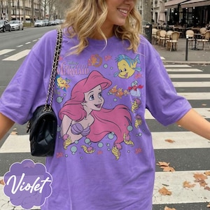 May include: A violet t-shirt with a graphic of Ariel from Disney's The Little Mermaid. The design includes Ariel with pink hair, fish, and bubbles. The shirt has short sleeves and a relaxed fit.