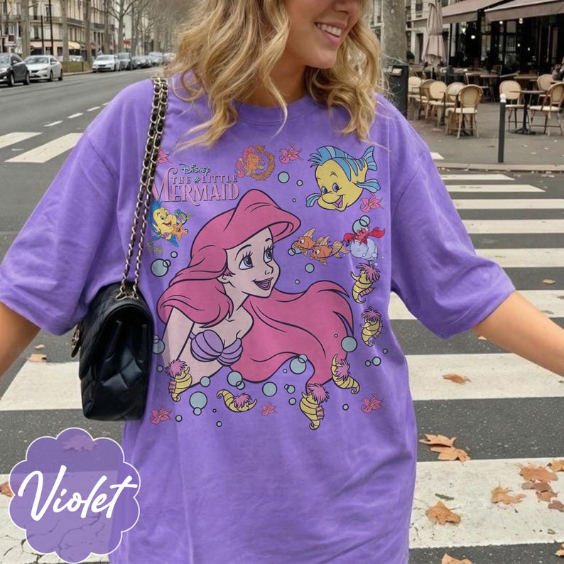 May include: A violet t-shirt with a graphic of Ariel from Disney's The Little Mermaid. The design includes Ariel with pink hair, fish, and bubbles. The shirt has short sleeves and a relaxed fit.