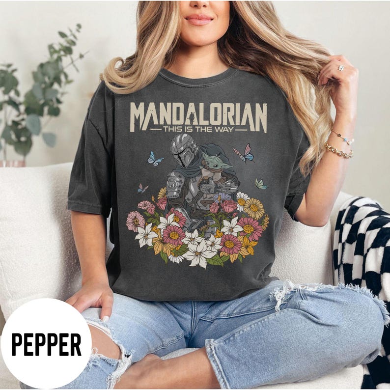 Puede incluir: Camiseta gris oscuro con el texto "MANDALORIAN - THIS IS THE WAY". El dise&ntilde;o muestra al Mandaloriano y a Grogu rodeados de flores y mariposas. Una prenda c&oacute;moda e informal.
