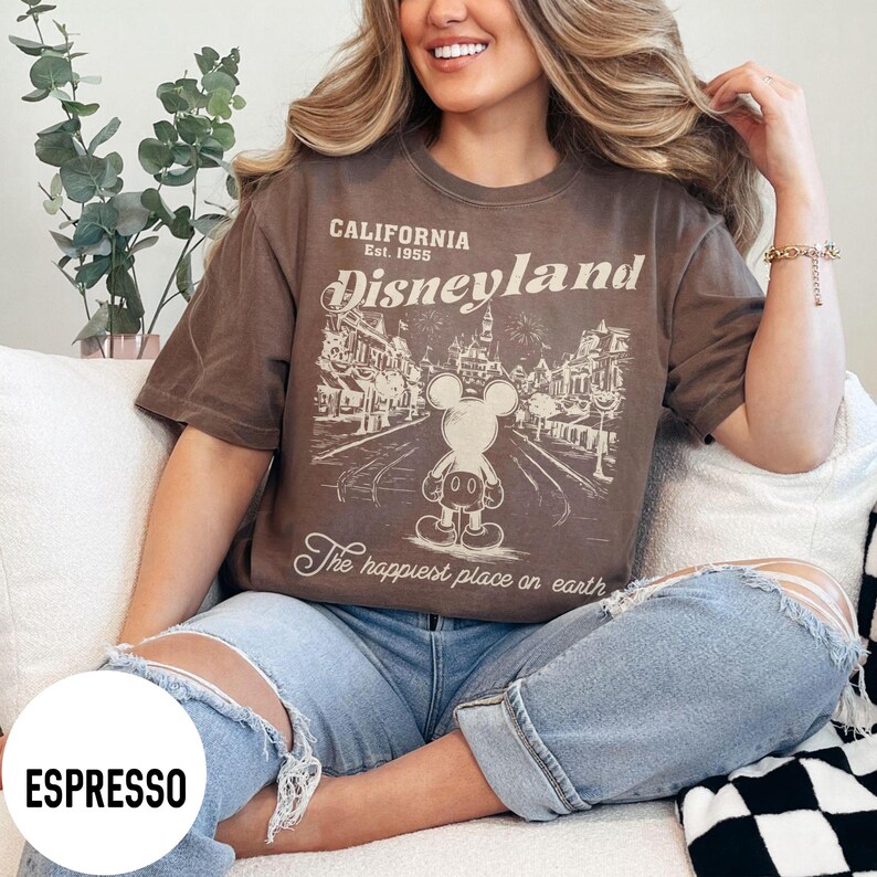 Może przedstawiać: Koszulka w kolorze espresso z grafiką Disneyland. Projekt przedstawia Myszka Miki przed sceną uliczną z napisem "CALIFORNIA Est. 1955 Disneyland" i "The happiest place on earth."