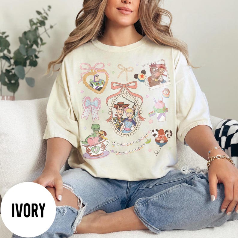 May include: Ivory-colored t-shirt featuring a collection of cartoon character graphics. The design includes images of a cowboy and cowgirl, a green dinosaur, and other characters. The word "IVORY" is printed in a white circle at the bottom.