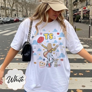 May include: A white t-shirt featuring a graphic print of a person with long hair playing a guitar, wearing a yellow plaid shirt and cow-print pants. The design includes blue clouds, a red hat, musical notes, and blocks with the letters TS.