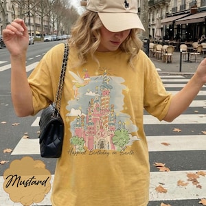 Puede incluir: Camiseta de color mostaza con una ilustraci&oacute;n colorida de un castillo, un hada y el texto "Happiest Birthday on Earth". La camiseta tiene mangas cortas y un corte holgado. La palabra "Mustard" est&aacute; escrita en cursiva en la parte inferior.