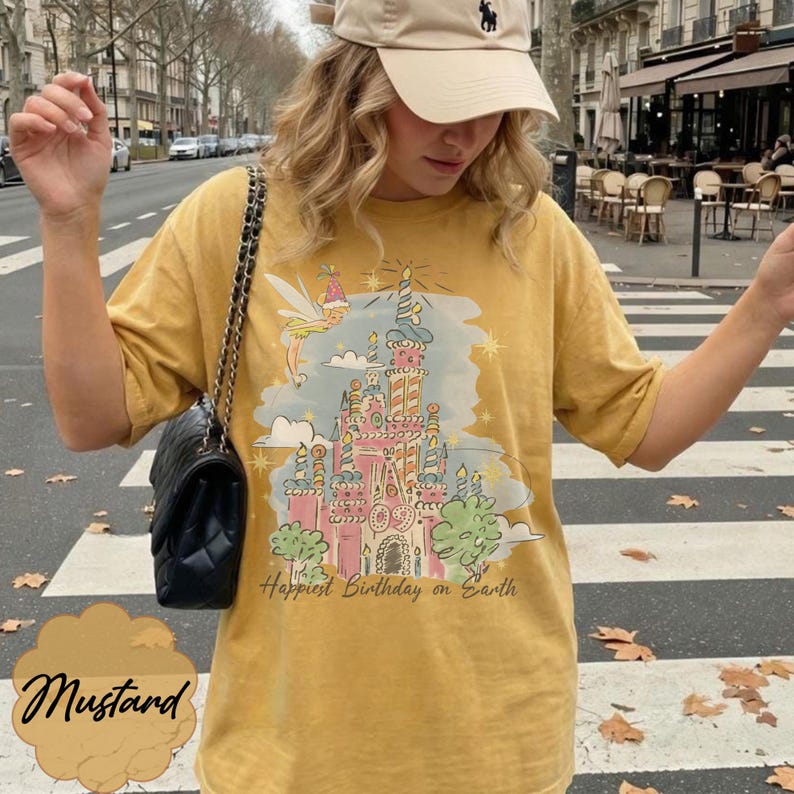 Puede incluir: Camiseta de color mostaza con una ilustraci&oacute;n colorida de un castillo, un hada y el texto "Happiest Birthday on Earth". La camiseta tiene mangas cortas y un corte holgado. La palabra "Mustard" est&aacute; escrita en cursiva en la parte inferior.