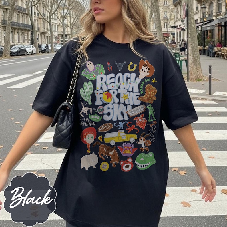 Pode incluir: Camiseta preta com um design gr&aacute;fico colorido com personagens e texto de um filme de anima&ccedil;&atilde;o popular. O texto na camiseta diz "REACH FOR THE SKY". A camiseta tem um corte casual e oversized.