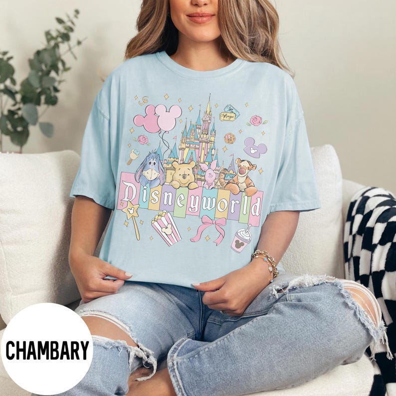 May include: Light blue t-shirt with a colorful "Disneyworld" graphic featuring cartoon characters, a castle, and other themed elements. The shirt has short sleeves and a relaxed fit.