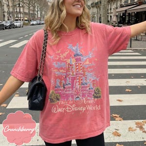 K&ouml;nnte beinhalten: Ein korallenfarbenes Kurzarm-T-Shirt mit einer Schlossgrafik und dem Text "25 This is Now! Walt Disney World 1997". Das T-Shirt hat einen Rundhalsausschnitt und eine lockere Passform, kombiniert mit einer schwarzen gesteppten Umh&auml;ngetasche.