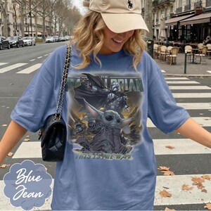 Puede incluir: Una camiseta azul con un estampado gr&aacute;fico de un personaje con armadura y una peque&ntilde;a criatura de orejas grandes. El texto dice THE MANDALORIAN y THIS IS THE WAY. La camiseta tiene mangas cortas y un cuello redondo.