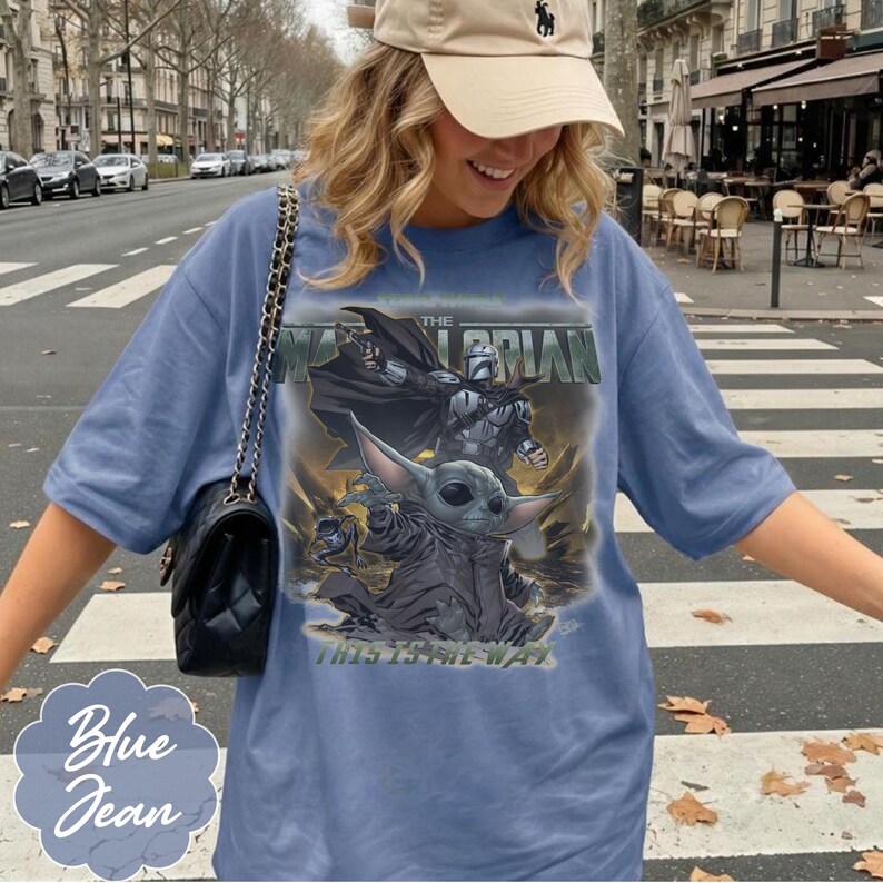 Puede incluir: Una camiseta azul con un estampado gr&aacute;fico de un personaje con armadura y una peque&ntilde;a criatura de orejas grandes. El texto dice THE MANDALORIAN y THIS IS THE WAY. La camiseta tiene mangas cortas y un cuello redondo.