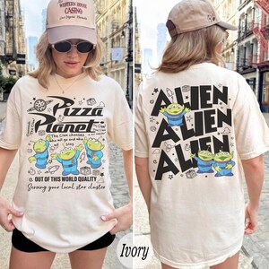 K&ouml;nnte beinhalten: Elfenbeinfarbenes Pizza Planet T-Shirt mit einer Grafik von drei gr&uuml;nen Aliens und dem Pizza Planet Logo auf der Vorderseite. Auf der R&uuml;ckseite des Shirts steht das Wort "ALIEN" wiederholt mit Alien-Grafiken.
