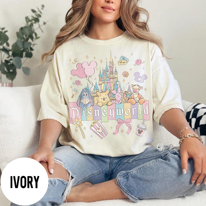 May include: Ivory-colored t-shirt featuring a colorful Disneyworld graphic. The design includes a castle, balloons, and various Disney characters like Winnie the Pooh and Tigger. The word "Disneyworld" is prominently displayed.