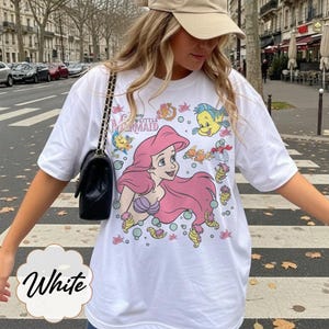 May include: White t-shirt with a graphic of Ariel from Disney's The Little Mermaid. The design includes Ariel with pink hair, Flounder, and other sea creatures. The shirt has the text "The Little Mermaid".