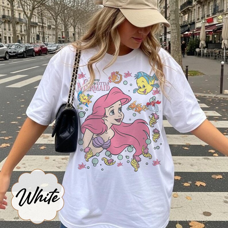 May include: White t-shirt with a graphic of Ariel from Disney's The Little Mermaid. The design includes Ariel with pink hair, Flounder, and other sea creatures. The shirt has the text "The Little Mermaid".