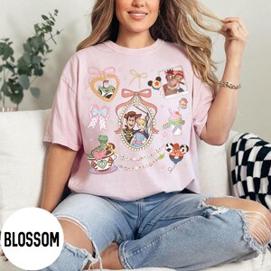 May include: A light pink t-shirt featuring various Toy Story character graphics. The design includes images of Buzz Lightyear, Woody, Bo Peep, and others, framed by bows and decorative elements. The word "BLOSSOM" is printed on the bottom left.