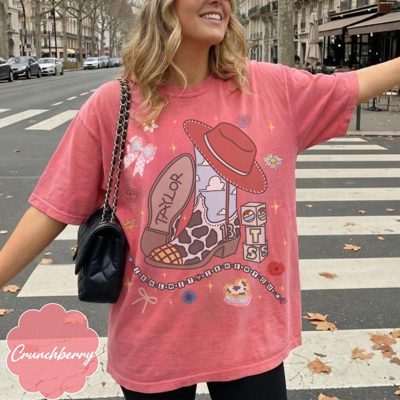 May include: A coral-colored short-sleeve t-shirt featuring a graphic print of a cowboy boot with a cow-print pattern, a red cowboy hat, a bow, and stacked blocks with the letters T, S, and the number 13. The text Taylor is on the boot.