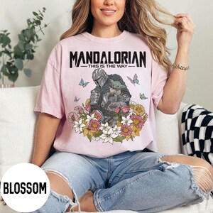 Puede incluir: Camiseta rosa claro con un dise&ntilde;o del Mandaloriano y Grogu, rodeados de flores y mariposas. El texto "MANDALORIAN - THIS IS THE WAY" est&aacute; impreso en negro en la parte superior.