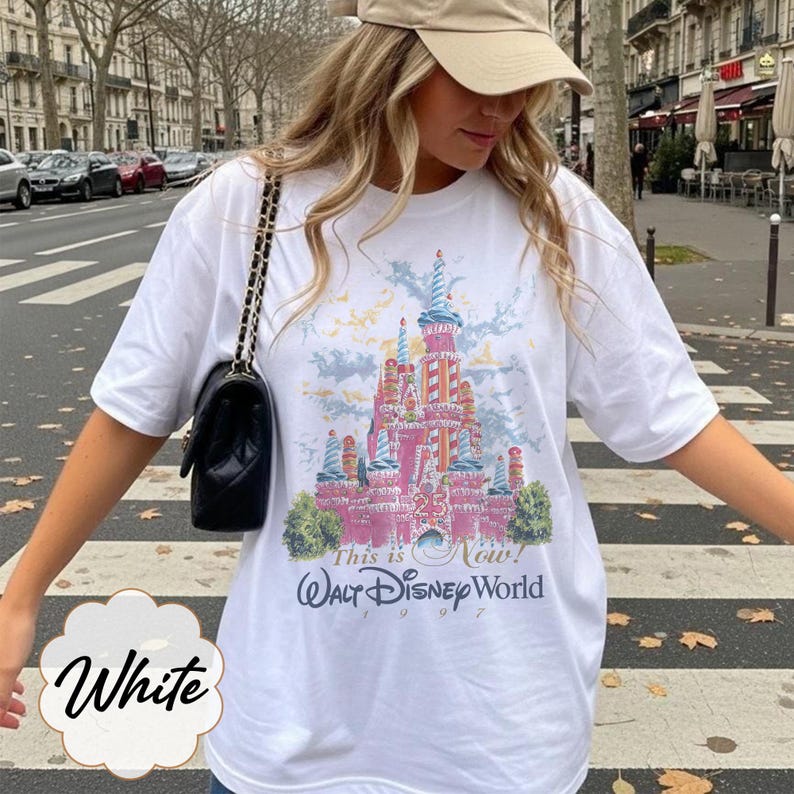 K&ouml;nnte beinhalten: Ein wei&szlig;es T-Shirt mit kurzen &Auml;rmeln und einer bunten Grafik eines Schlosses mit zuckerstangenartigen T&uuml;rmen. Der Text lautet This is Now! Walt Disney World 1997. Das Design zeigt gr&uuml;ne B&uuml;sche an der Basis und einen hellblauen Himmel.