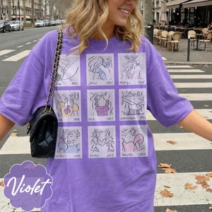 May include: A violet-colored t-shirt featuring a grid of cartoon sketches with dates. The sketches appear to be of animated characters. The word "Violet" is written in a cursive font at the bottom left of the shirt.