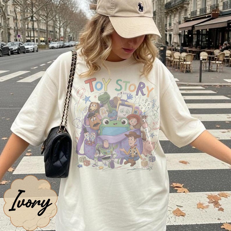 May include: An ivory short-sleeved t-shirt featuring a colourful graphic print of Toy Story characters including Woody, Buzz Lightyear, and a green frog. The text Toy Story is printed in multicoloured letters above the characters on the front.