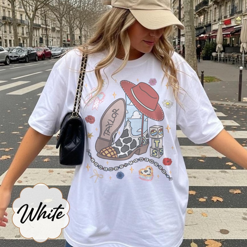 May include: A white short-sleeve t-shirt featuring a graphic print of a cowboy boot with a cow print pattern, a red hat, and stacked blocks with the letters T, S, and S. The design includes the name Taylor, small flowers, and stars.