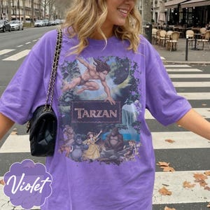 Op de afbeelding: Een violetkleurig T-shirt met een Tarzan-afbeelding. De afbeelding bevat het woord "Tarzan" in een bruin lettertype, samen met illustraties van Tarzan en andere personages uit de film. Een zwarte handtas met een kettingriem is zichtbaar.