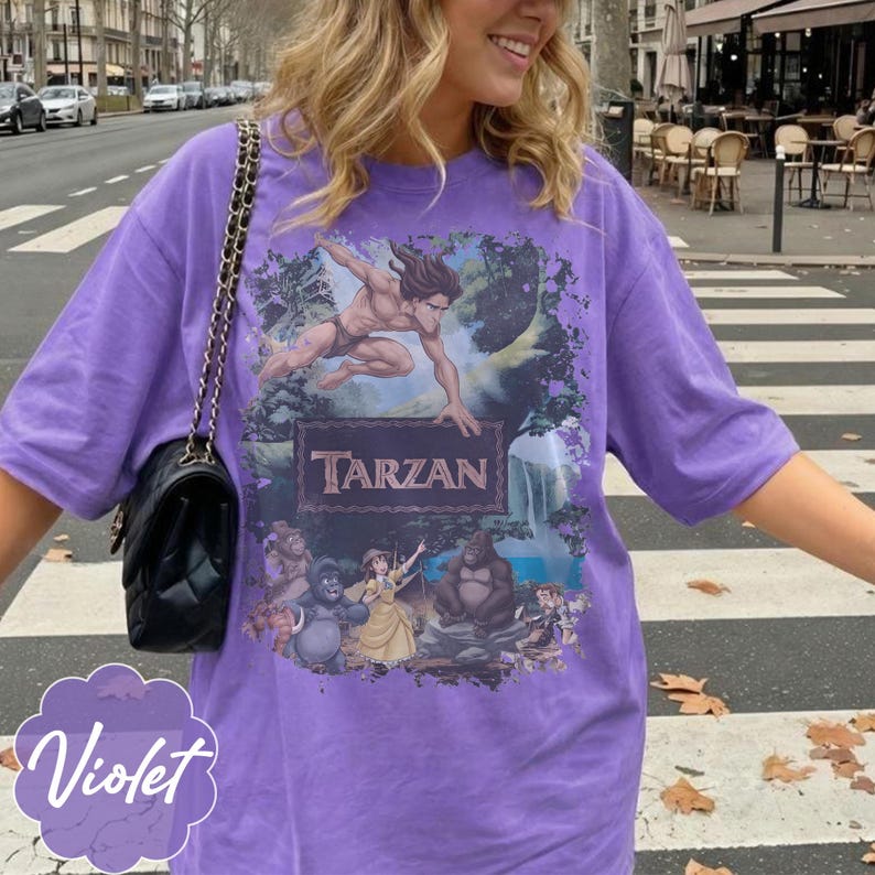 Op de afbeelding: Een violetkleurig T-shirt met een Tarzan-afbeelding. De afbeelding bevat het woord "Tarzan" in een bruin lettertype, samen met illustraties van Tarzan en andere personages uit de film. Een zwarte handtas met een kettingriem is zichtbaar.