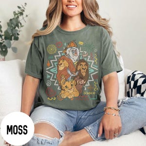 Puede incluir: Camiseta verde musgo con un gr&aacute;fico colorido de personajes de El Rey Le&oacute;n. El dise&ntilde;o incluye a Simba, Mufasa, Scar, Tim&oacute;n, Pumba y Zaz&uacute;, con patrones de inspiraci&oacute;n tribal. La camiseta es de corte holgado.