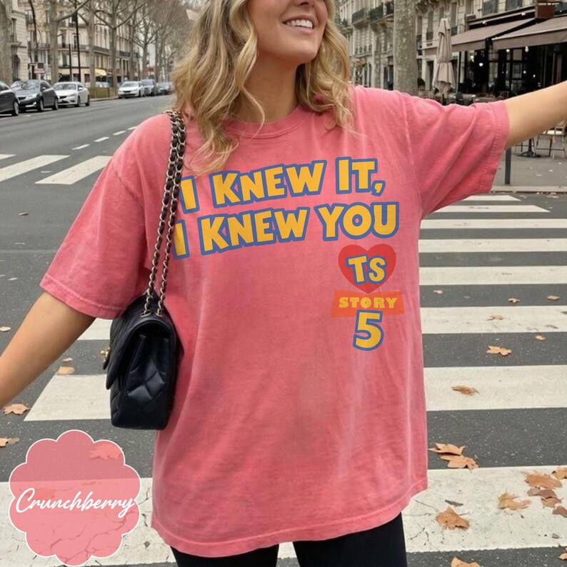 May include: A coral-colored short-sleeve t-shirt featuring the yellow text I KNEW IT, I KNEW YOU with a red heart containing the letters TS, the word STORY, and the number 5 printed in blue and yellow on the front of the garment.